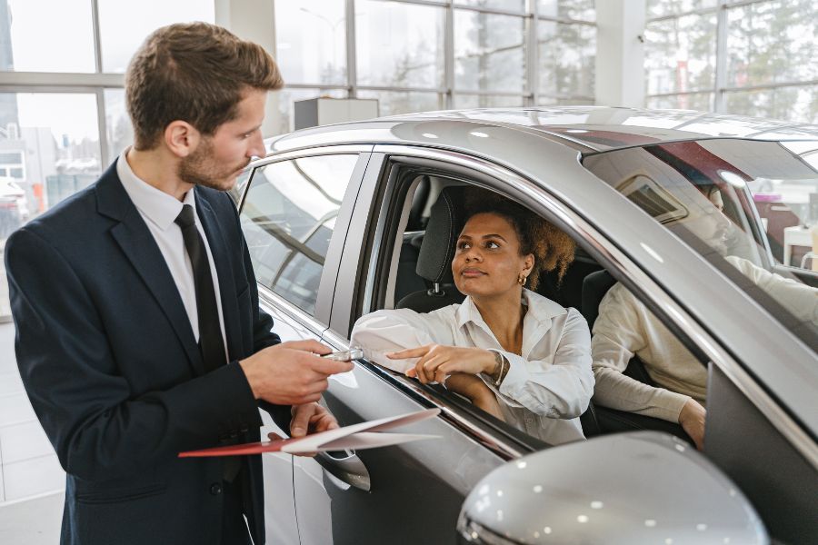 An employee is standing in front of a car with a clipboard in his hand. A woman is sitting in the vehicle. A symbolic image for the vehicle valuation