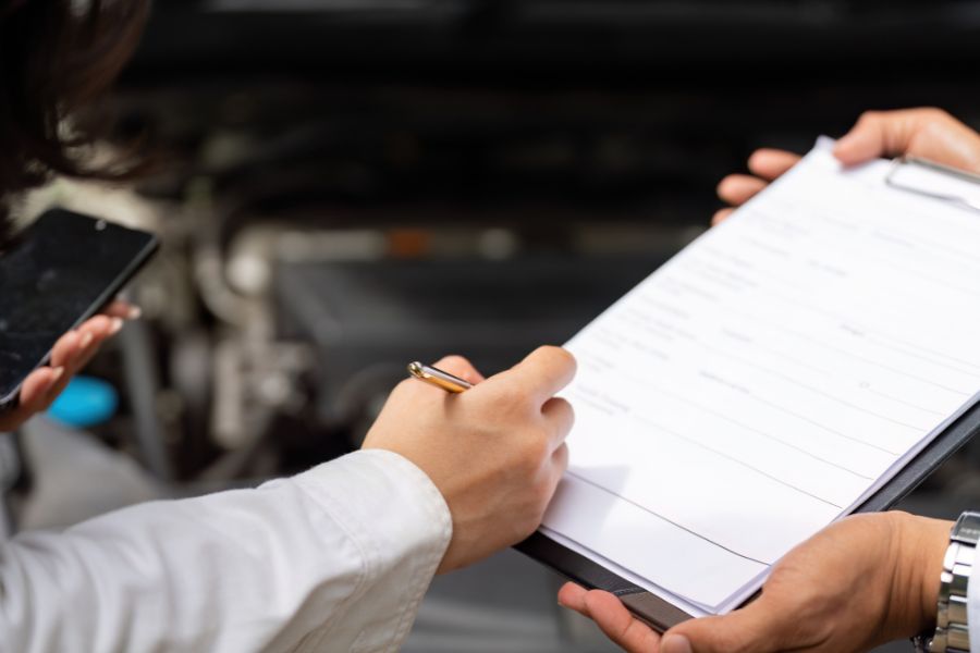 A man signs a paper on a clipboard in front of a car. A symbolic image for the vehicle registration document.