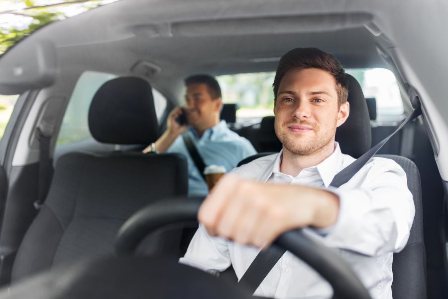 A man in a white shirt is driving a car. Another man is sitting in the back seat. A symbolic image for drivers.