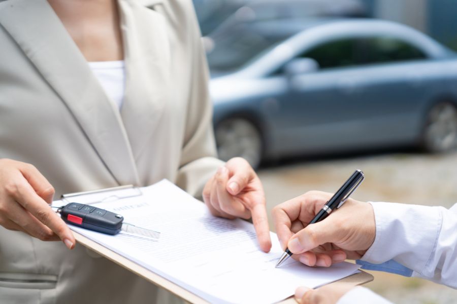 A man signs his name on a clipboard. There is a car key on the clipboard. A symbolic image for finance leasing.