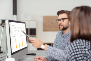 An employee explains an evaluation for fleet optimization with the Fleet+ software to a colleague on the computer. The graphic shows bar charts on vehicle usage. Carano supports companies with digital solutions for efficient, sustainable and transparent fleet management as part of modern mobility strategies.