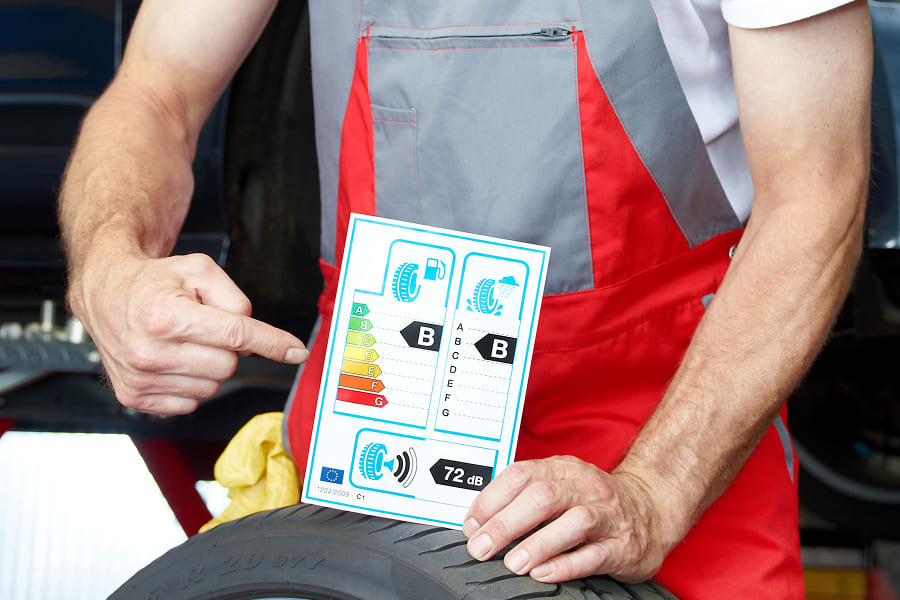 A mechanic holds a vehicle label with information on efficiency class, fuel consumption, CO₂ emissions and noise level next to a car tire. The image illustrates the label for assessing the environmental impact and operating costs of a vehicle and supports transparent vehicle selection in fleet management.